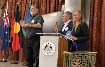A smiling Terry Hartmann holding the Biometrics Institute’s Lifetime Achievement Award alongside Biometrics Institute Chairman, Jakob Glynstrup and Institute CEO, Isabelle Moeller.