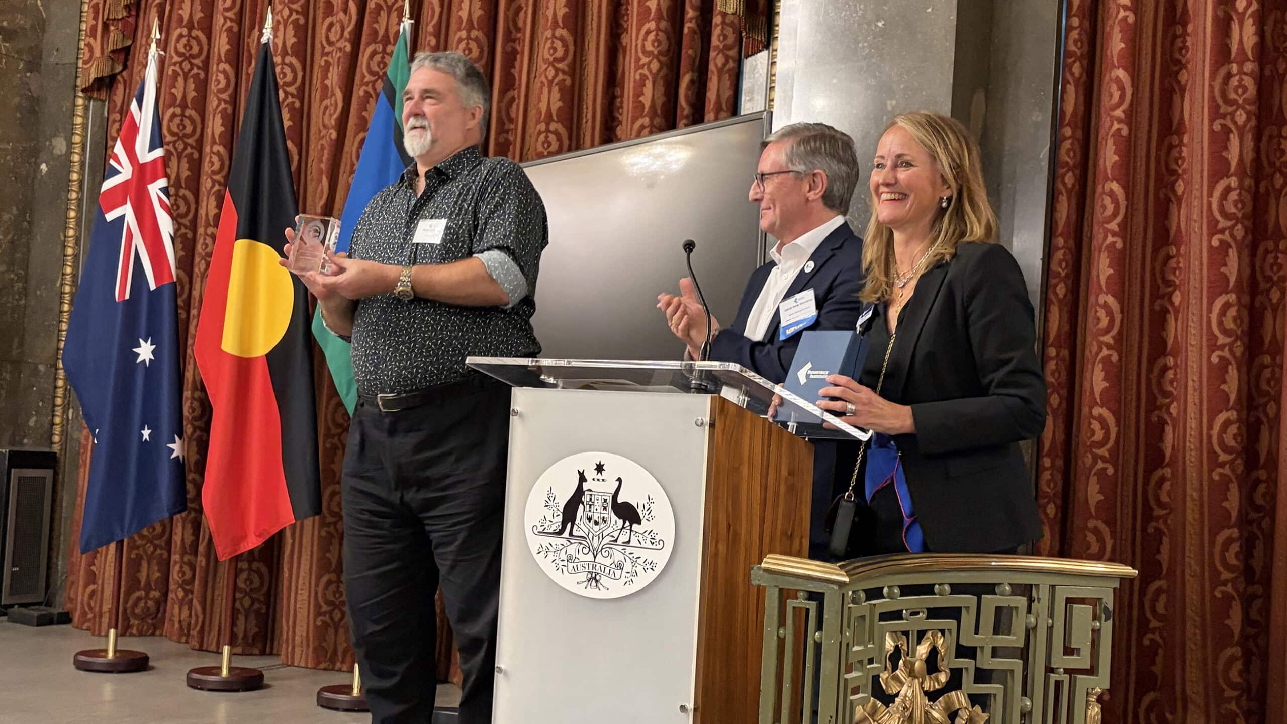 A smiling Terry Hartmann holding the Biometrics Institute’s Lifetime Achievement Award alongside Biometrics Institute Chairman, Jakob Glynstrup and Institute CEO, Isabelle Moeller.